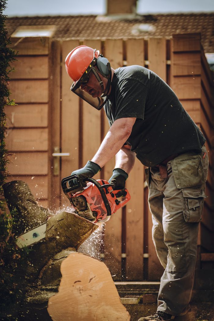 Worker using a chainsaw to cut wood, wearing safety gear in an outdoor setting.